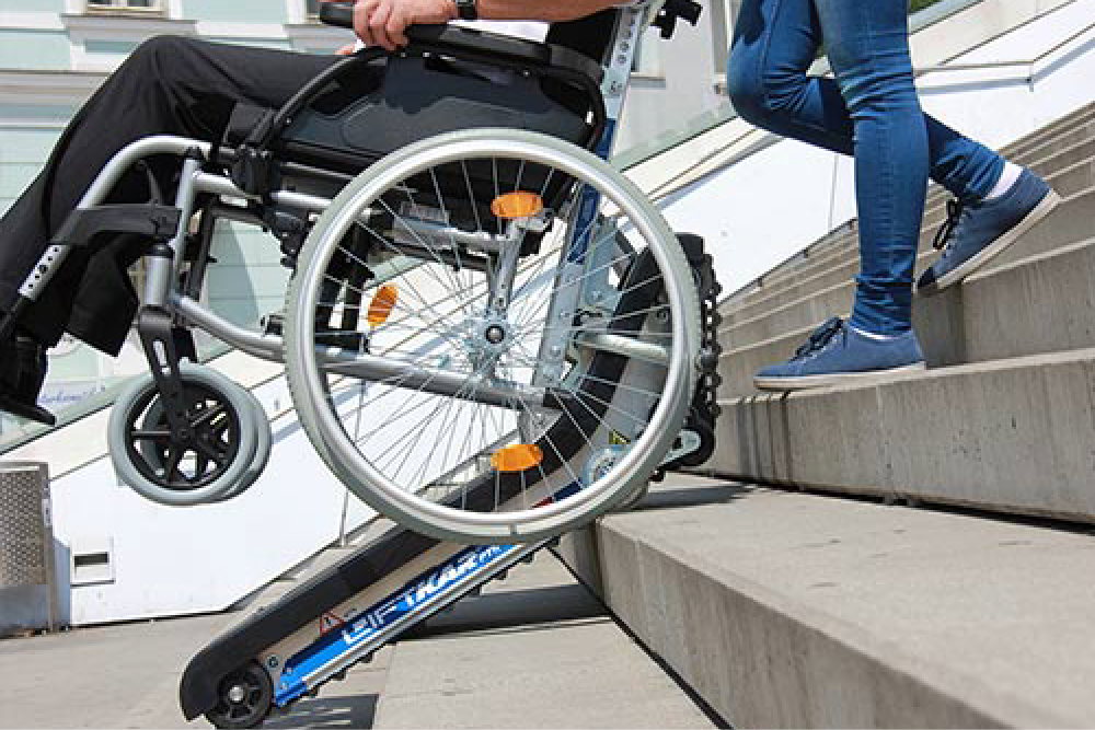 Osoba niepełnosprawna w asyście drugiej osoby, zjeżdża po schodach na schodołazie.